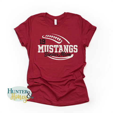 A red short-sleeve crewneck t-shirt with a two-color white and black glitter football with the text of Mustangs Senior Mom running in the middle of the football.