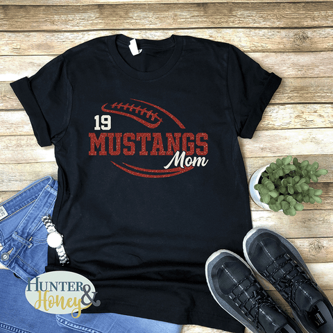 A black short-sleeve crewneck t-shirt with a two-color red and white glitter football with the text of Mustangs Mom running in the middle of the football.