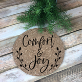 7" round natural cork trivet with a black graphic that reads Comfort and Joy in a script with leaves flanking both sides of the graphic. 