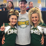 A dad wearing a Damascus Hornets Varsity Cheer Dad gray tee with his two daughters at a cheer competition.