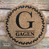 7" round natural cork customized trivet with a single large letter monogram and family last name under the letter surrounded by a laurel border.