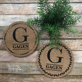 A 2-piece set of 7" round natural cork customized trivets with a single large letter monogram and family last name under the letter surrounded by a leaf and dot or a laurel border.