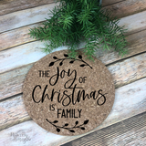 7" round natural cork trivet with black script "The Joy of Christmas is Family" graphic flanked by leaves.