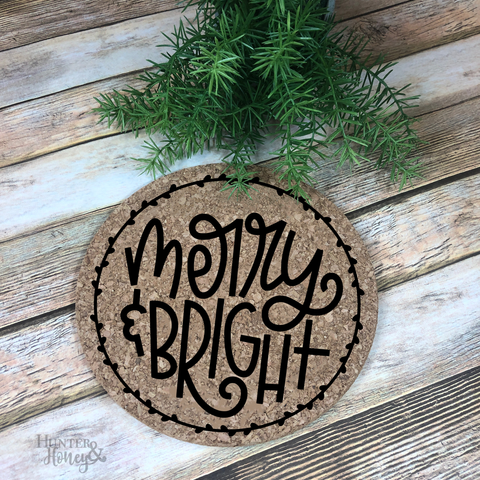 7" round natural cork trivet with a playful black script and printing Merry & Bright graphic surrounded by laurel border.