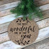 A 7" round natural cork trivet with black script graphic saying "The Most Wonderful Time of the Year"  and leaves gracing the top and bottom.